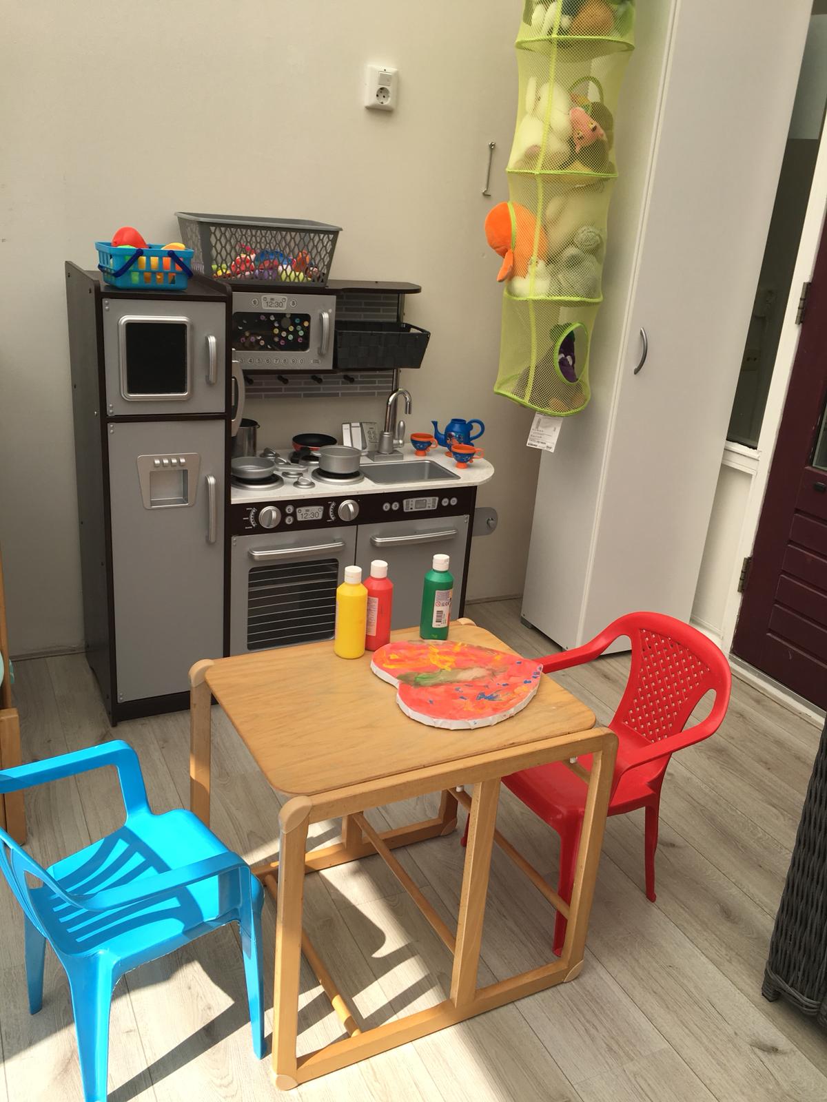 Kindertafel met potten verf en een geverfd hart, met een blauw en rood stoeltje en een speelkeuken
