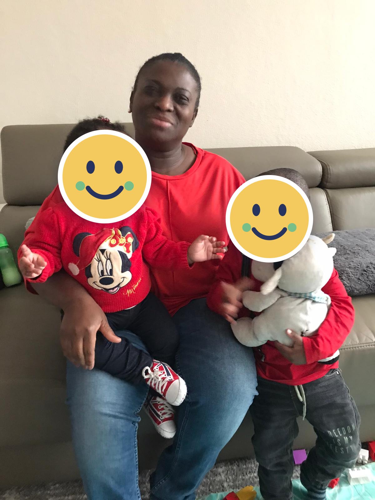 Een vrouw in een rode trui zit op de bank met een peuter op schoot en een jongetje met een knuffel naast zich; de gezichten van de kinderen zijn afgedekt met een smiley